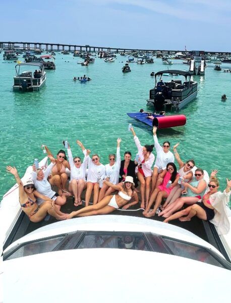 Group of friends lounging on a boat bow at a lively boat party in clear turquoise coastal waters with many anchored boats and a low bridge in the background on a sunny day