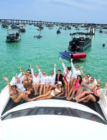 Group of friends lounging on a boat bow at a lively boat party in clear turquoise coastal waters with many anchored boats and a low bridge in the background on a sunny day