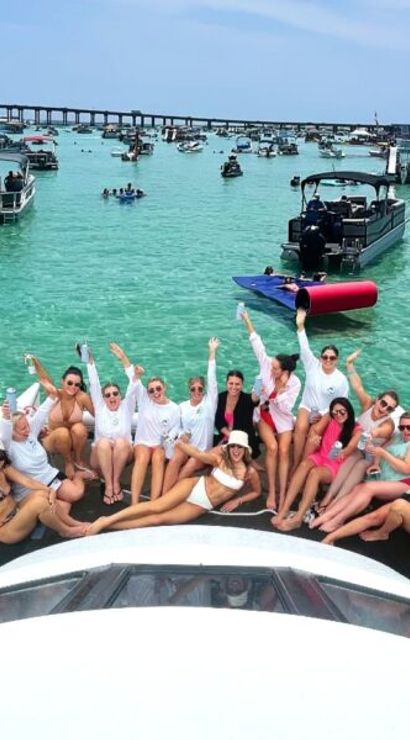 Group of friends lounging on a boat bow at a lively boat party in clear turquoise coastal waters with many anchored boats and a low bridge in the background on a sunny day