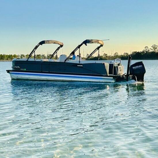 Pontoon boat with dual folding bimini tops and outboard motor cruising on calm blue water near a marina at golden hour.