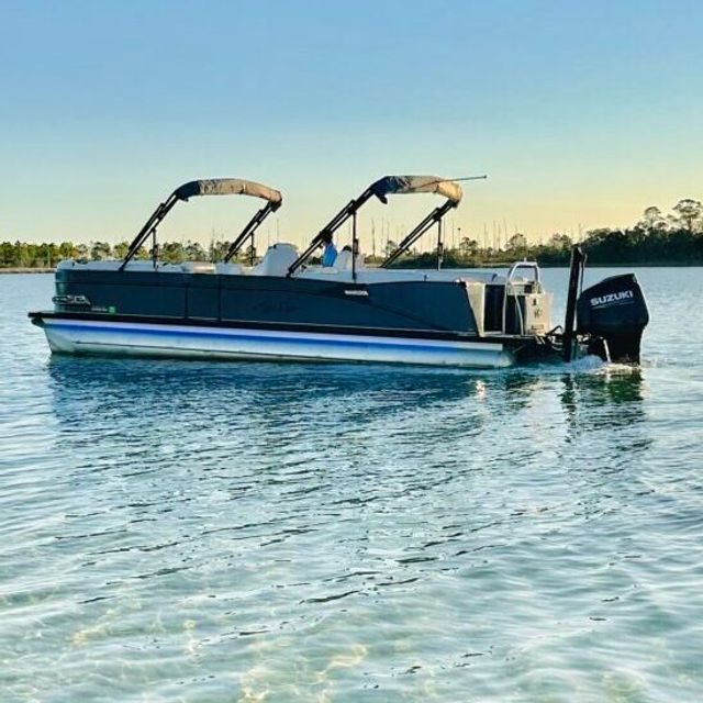 Pontoon boat with dual folding bimini tops and outboard motor cruising on calm blue water near a marina at golden hour.