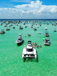 Aerial view of dozens of boats anchored at a crowded tropical sandbar in clear turquoise water, with a white catamaran in the foreground and people swimming and lounging under a blue sky with puffy clouds.
