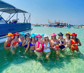 Group of women in colorful bikinis and cowboy hats waist-deep in clear turquoise water next to a pontoon boat, holding drinks at a sunny sandbar party with floating bar and boats in the background.