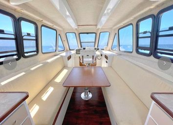 Sunny yacht cabin interior with wraparound windows, cream cushioned bench seating, central wood table, helm station forward and clear ocean views