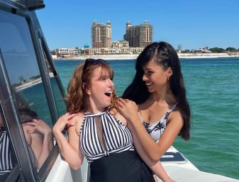 Two friends laughing on a boat in clear turquoise coastal water with a sandy beach and beachfront high-rise towers under a bright blue sky
