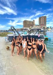Friends posing on a sandy shore beside a docked pontoon boat, all in swimsuits and sunglasses, with a small dog in a life jacket nearby and blue sky and waterfront resort buildings in the background.