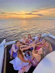 Celebratory group of women wearing sashes toasting with drinks on a catamaran at golden sunset over calm ocean waters