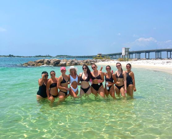 Nine women in swimsuits posing waist-deep in clear turquoise water on a sunny beach, holding drinks with a rocky jetty and a long bridge stretching across the shoreline behind them.