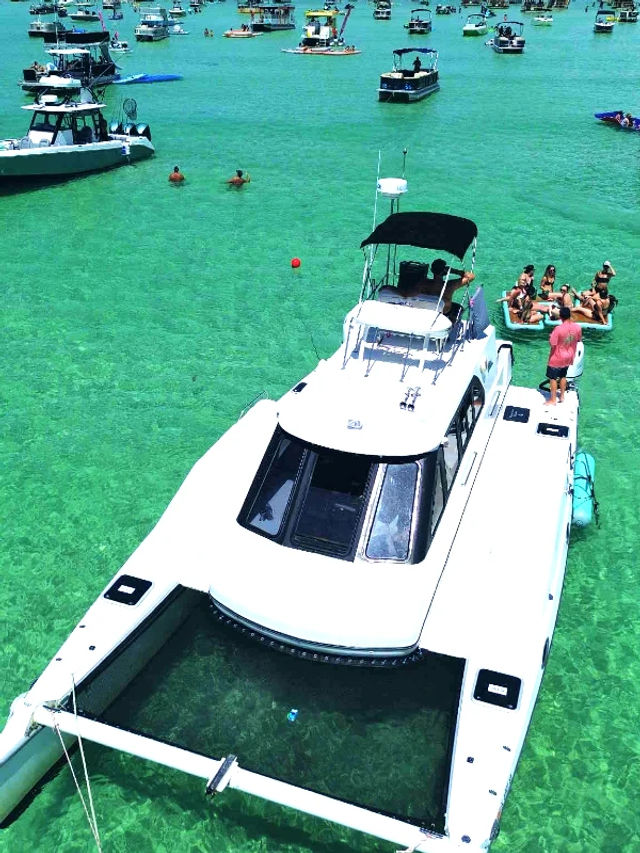 White catamaran anchored in crystal-clear turquoise tropical water on a sunny day, people lounging on an inflatable raft alongside and dozens of boats anchored in the background