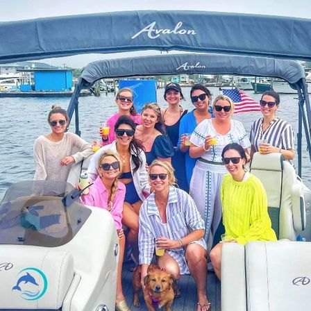 Group of women on a pontoon boat at a marina, smiling and holding drinks for a daytime boat party, small brown dog at their feet and an American flag fluttering in the background
