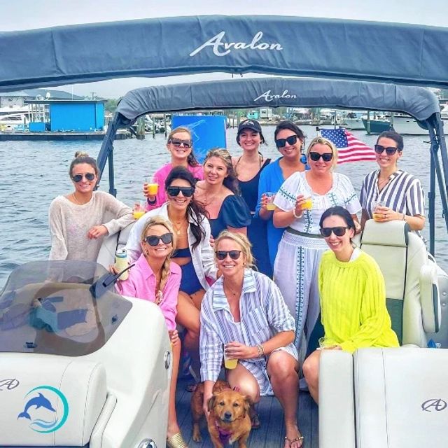Group of women on a pontoon boat at a marina, smiling and holding drinks for a daytime boat party, small brown dog at their feet and an American flag fluttering in the background