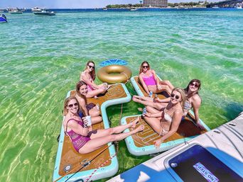 Six smiling women lounging on connected paddleboard-style floats in clear green water, sipping drinks near anchored boats on a sunny summer bay.