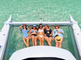 Five friends in swimsuits lounging on a catamaran trampoline over clear turquoise water, sunbathing and laughing on a tropical boat trip.