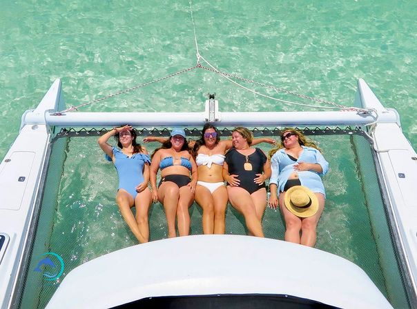 Five friends in swimsuits lounging on a catamaran trampoline over clear turquoise water, sunbathing and laughing on a tropical boat trip.