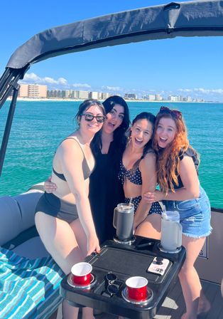 Four smiling friends in swimsuits posing on a pontoon boat with turquoise water, beachfront high-rise condos on the horizon and clear blue sky on a sunny coastal vacation.