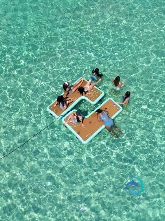 Aerial view of friends in swimsuits lounging on and swimming around a tethered modular floating platform in crystal-clear turquoise tropical water on a sunny day