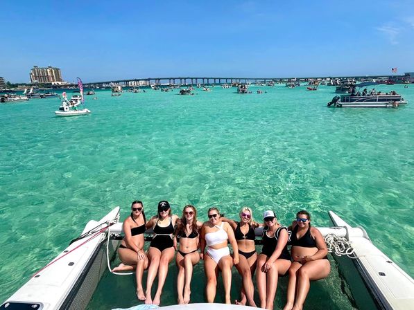 Group of seven friends in swimsuits lounging on a catamaran net over crystal-clear turquoise bay, surrounded by anchored boats and a low coastal bridge under a bright blue sky.