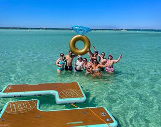 Cheerful group of beachgoers waist-deep in crystal-clear turquoise water, posing with a giant gold inflatable ring topped like a diamond and celebrating near mint-trim floating dock platforms under a bright blue sky.