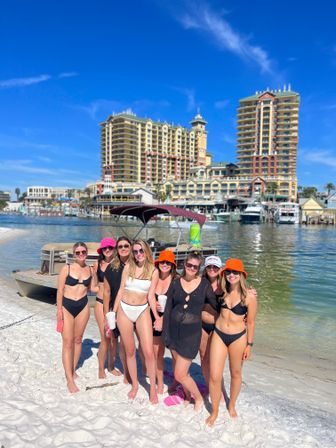 Seven women in swimsuits and sun hats posing on a white-sand beach by a docked boat and marina, with colorful high-rise beachfront resort buildings and a clear blue sky on a sunny vacation day.