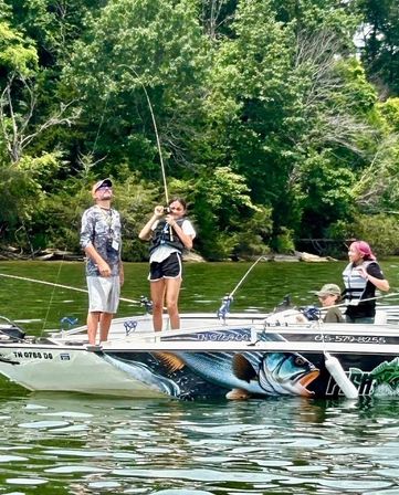 Four anglers in life jackets on a painted fishing boat reeling in a catch on a tree-lined lake — decorative bass graphic on the hull, summer fishing scene.