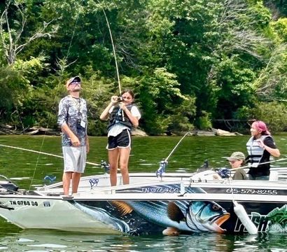Four anglers in life jackets on a painted fishing boat reeling in a catch on a tree-lined lake — decorative bass graphic on the hull, summer fishing scene.