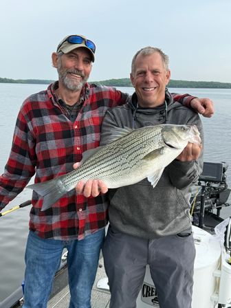 Two smiling anglers on a boat proudly holding a large striped bass over a calm freshwater lake — a big lake fishing catch.
