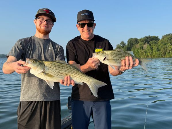 Two anglers on a fishing boat holding large striped bass over calm lake water at golden-hour with a tree-lined shoreline in the background, recreational lake fishing scene.