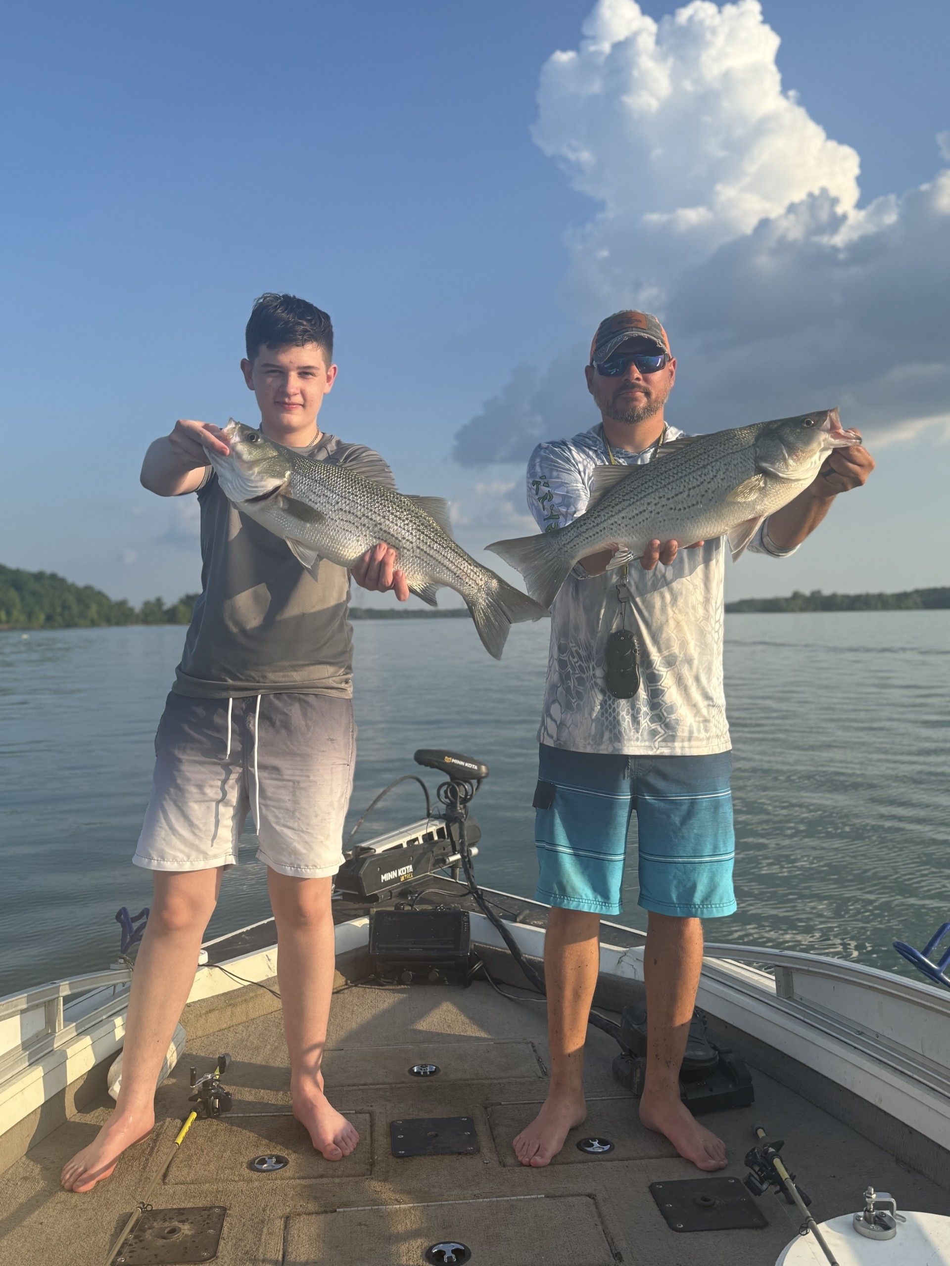 Two anglers barefoot on a fishing boat holding large striped bass over a calm freshwater lake under a blue sky with puffy clouds — proud summer catch