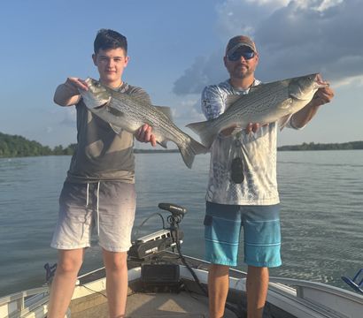 Two anglers barefoot on a fishing boat holding large striped bass over a calm freshwater lake under a blue sky with puffy clouds — proud summer catch