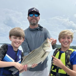 Smiling man and two boys in life jackets holding a large striped bass on a boat under a cloudy sky during a coastal fishing trip.