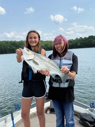 Two smiling people on a fishing boat at a tree-lined lake holding a large striped fish while wearing life jackets under a blue sky