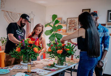 Four adults arranging vibrant orange-rose and purple-flower bouquets on a craft table during a DIY floral workshop in a cozy studio with a fiddle-leaf plant and artwork on the walls.