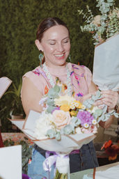 Smiling florist arranging a pastel bouquet of roses, eucalyptus, yellow daisies and purple blooms at a flower stall with a lush green backdrop