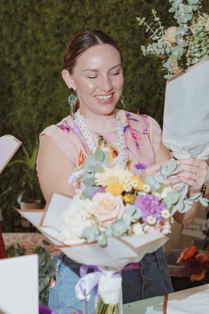 Smiling florist arranging a pastel bouquet of roses, eucalyptus, yellow daisies and purple blooms at a flower stall with a lush green backdrop