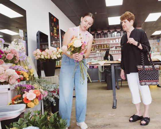 Florist arranging a mixed rose bouquet for a customer inside a bright local flower shop studio, surrounded by buckets of colorful roses, gerbera daisies and greenery.