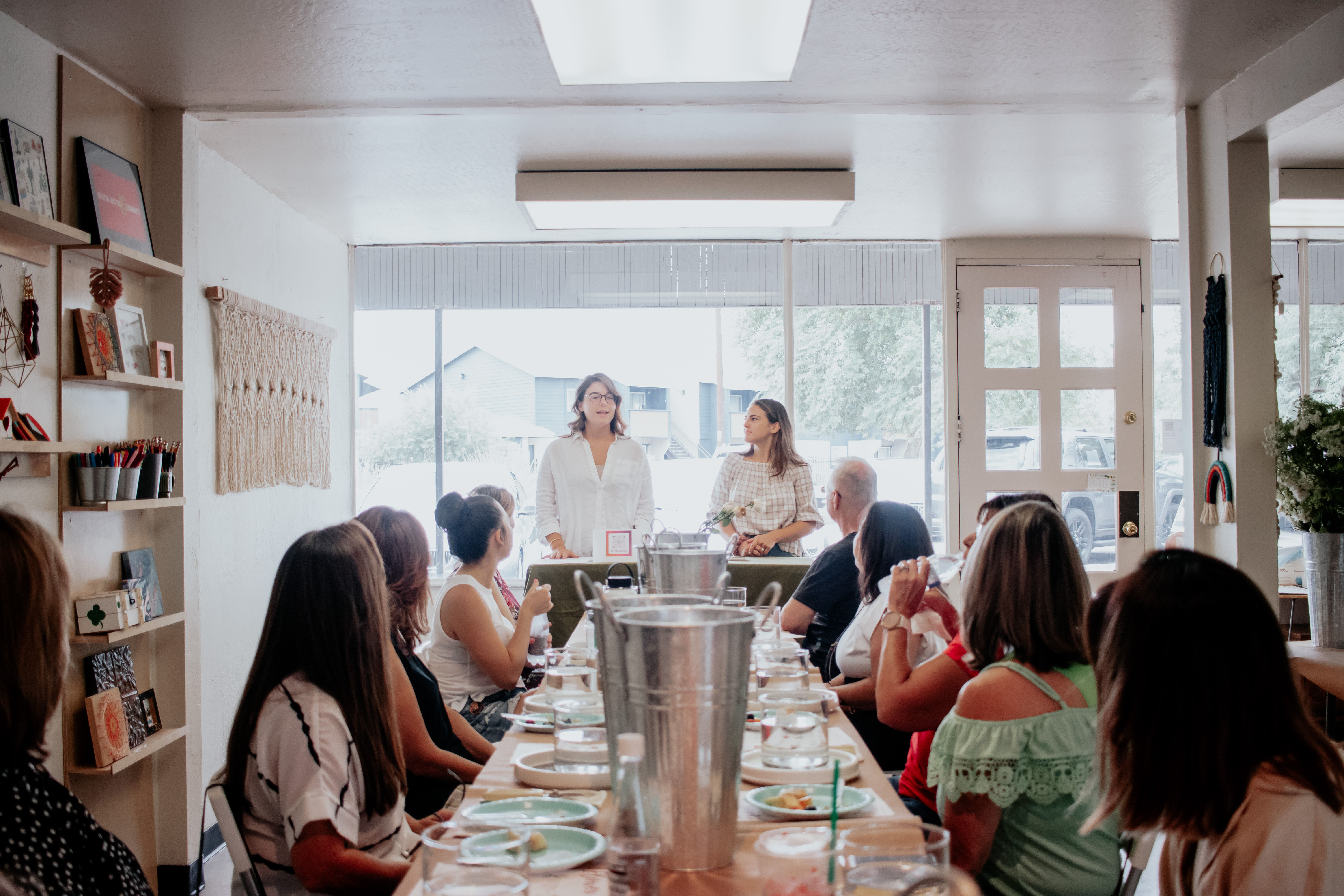 Two women presenting to a group of adults seated around a long table set with plates and drinks in a cozy sunlit community room with large front windows — casual workshop or luncheon.
