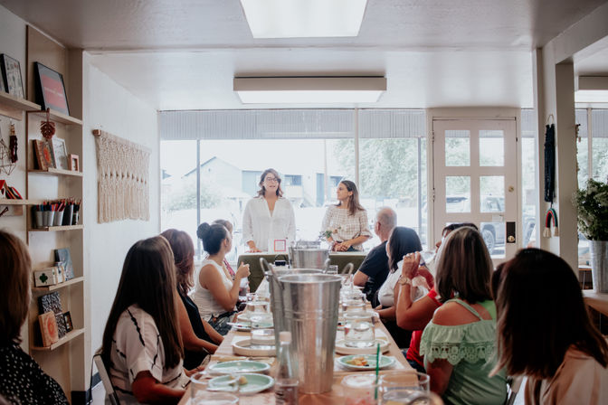 Two women presenting to a group of adults seated around a long table set with plates and drinks in a cozy sunlit community room with large front windows — casual workshop or luncheon.