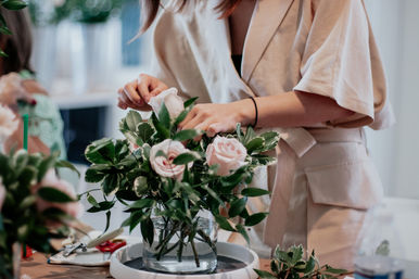 Hands arranging pale pink roses and variegated greenery in a glass vase on a workshop table — fresh bouquet styling indoors