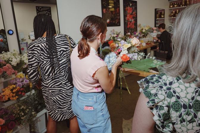 Hands-on floral workshop: people arranging a colorful bouquet in a bright studio surrounded by roses, hydrangeas and mixed greenery.