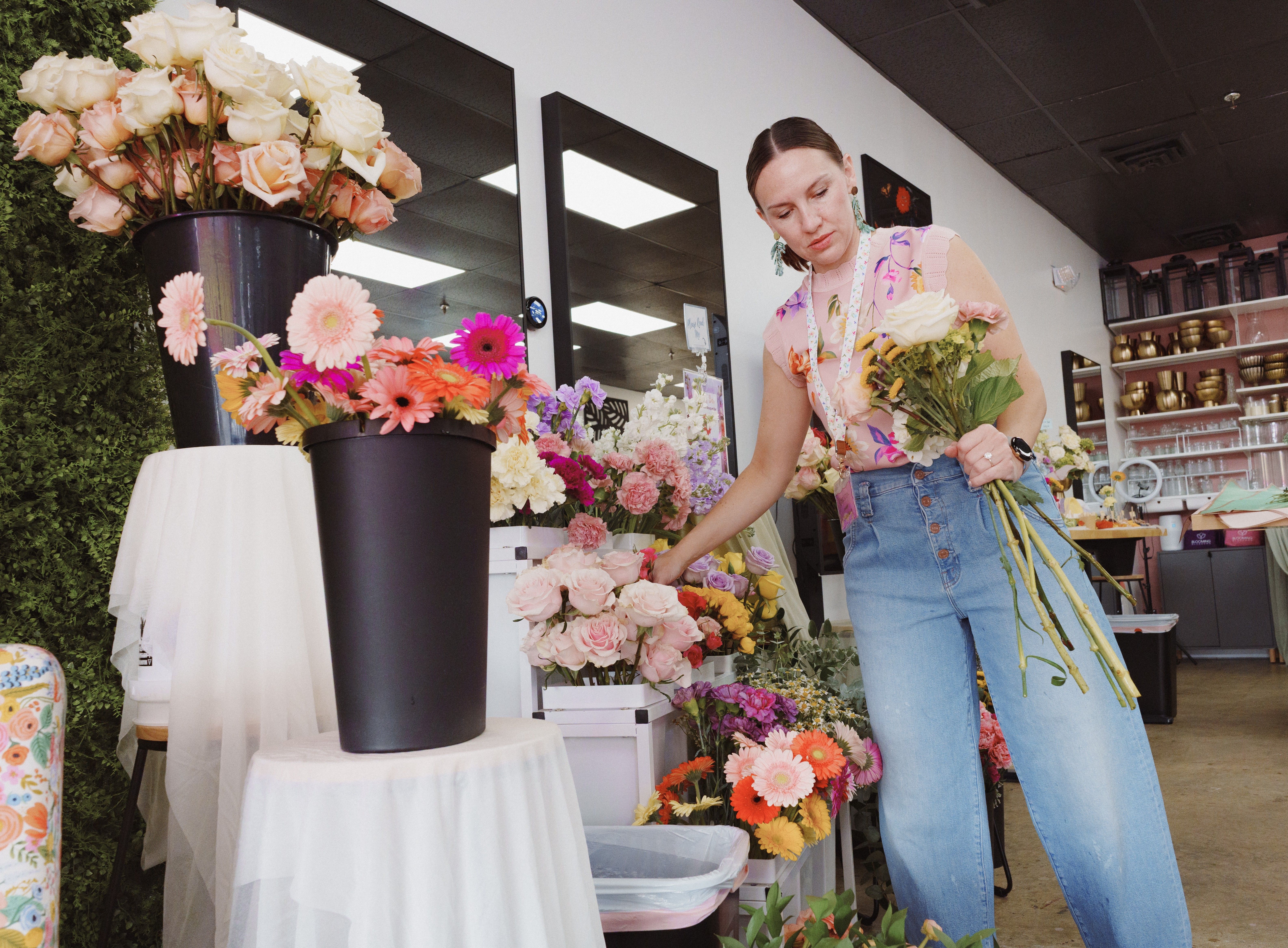 Florist arranging pastel roses and vibrant gerbera daisies in buckets inside a bright flower shop, holding a hand-tied bouquet while selecting stems for an arrangement.