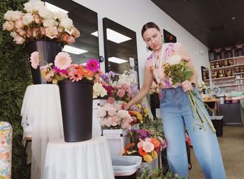 Florist arranging pastel roses and vibrant gerbera daisies in buckets inside a bright flower shop, holding a hand-tied bouquet while selecting stems for an arrangement.