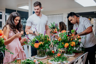 Adults in a hands-on indoor flower arranging class arranging vibrant orange roses, purple accents and lush greenery on a communal table