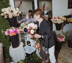 Shopper arranging bouquets in a bright indoor flower shop, selecting pink and orange gerbera daisies and roses from black display buckets — boutique florist scene