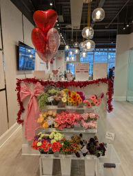 Valentine’s-themed flower display in a modern boutique florist with heart balloons, pink bow and red garland over shelves of roses, gerbera and mixed blooms by a storefront window