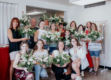 Smiling group in a bright studio holding homemade floral arrangements of pale pink roses and mixed greenery after an indoor flower-arranging workshop