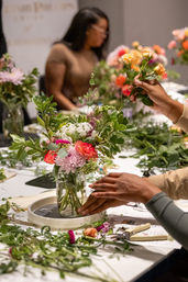 Hands arranging a colorful bouquet in a glass jar during an indoor flower-arranging workshop, with roses, chrysanthemums, greenery and clippers on the table.