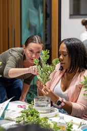 Hands-on floral workshop — two women arranging variegated green stems over a water jar as one guides the other at a busy indoor craft table.