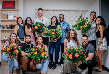 Happy, diverse group in a bright indoor floral workshop proudly holding vibrant orange-rose bouquets from a flower-arranging class.