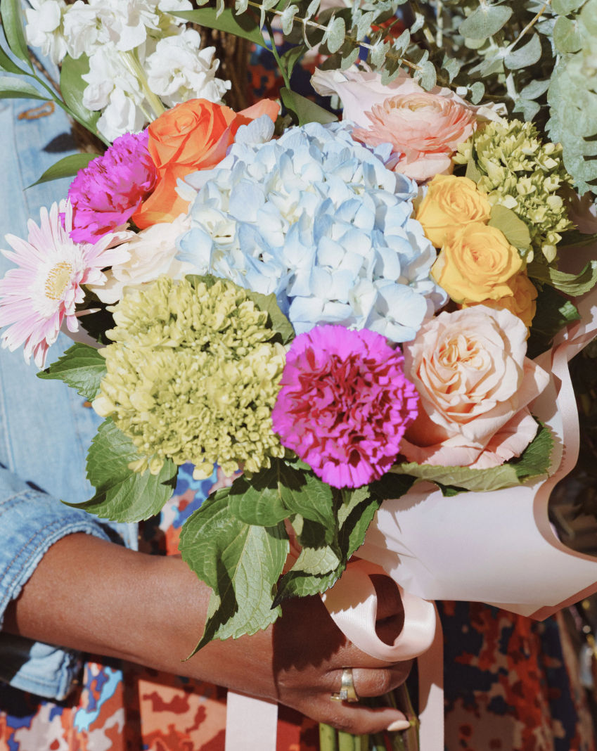 Close-up of a hand holding a vibrant mixed bouquet with blue hydrangea, peach and yellow roses, bright pink carnation, pale pink gerbera, green viburnum, eucalyptus sprigs and a pink satin ribbon