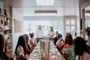 Two women presenting to a group of adults seated around a long table set with plates and drinks in a cozy sunlit community room with large front windows — casual workshop or luncheon.